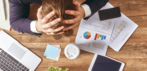 A solopreneur is holding his head while sitting at a desk with papers and a laptop, seeking ways to increase productivity.