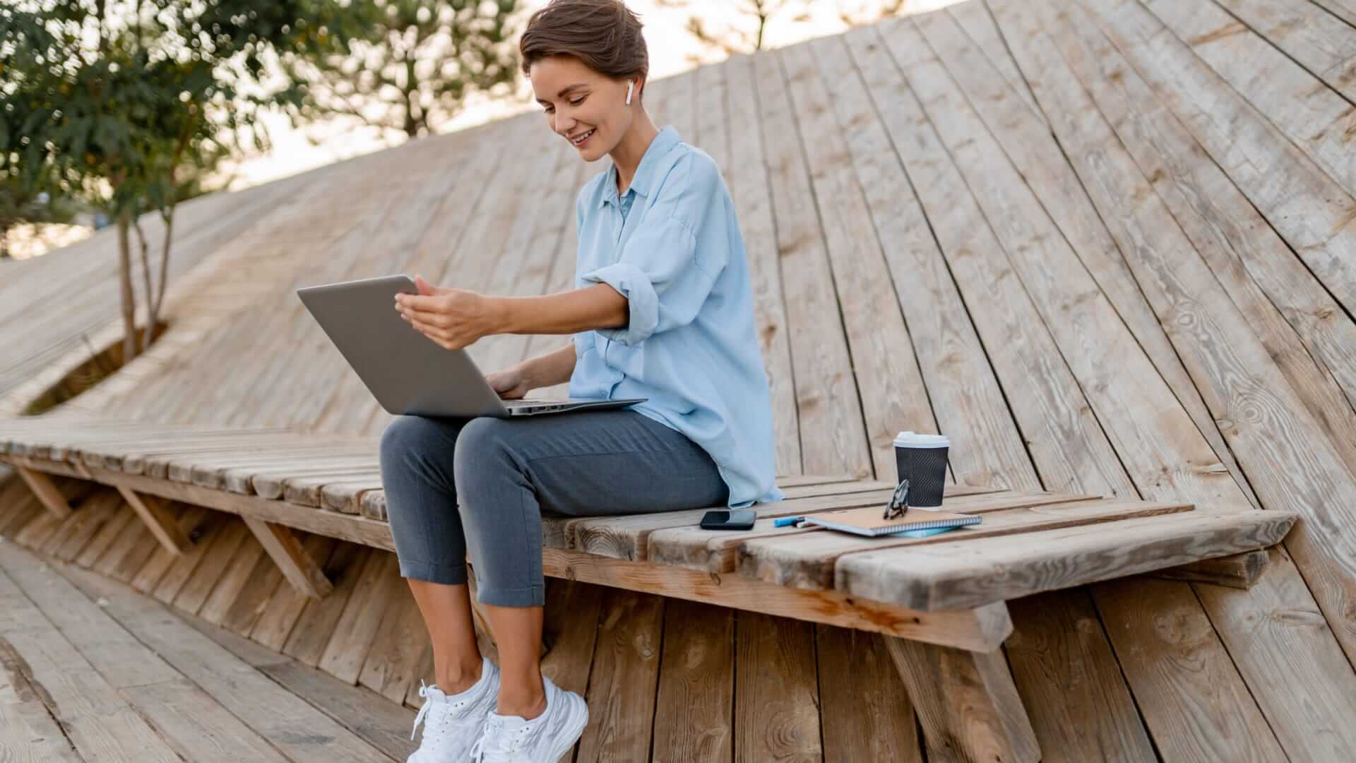 A woman sitting on a wooden bench using the latest employee timesheet app on her laptop.