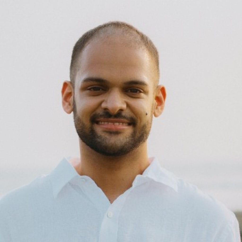 Ranjit Bhinge, a man with a light beard smiling at the camera, wearing a light blue shirt with a natural backdrop.