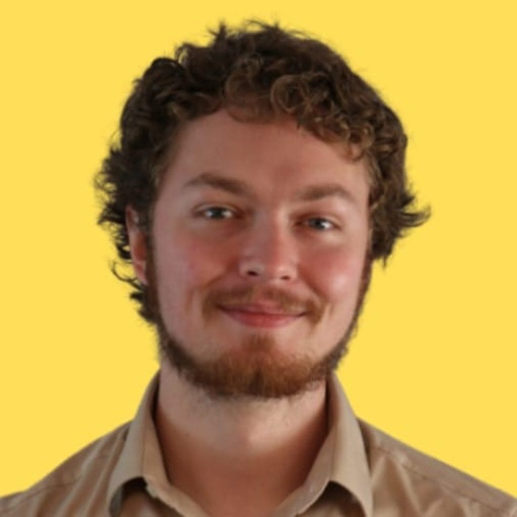 A smiling young man named Nick Baird with curly hair and a light beard on a yellow background. He is wearing a brown shirt.