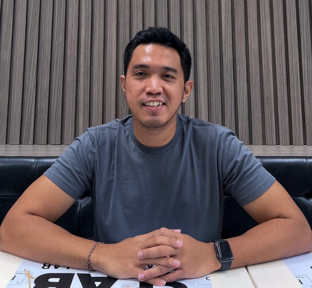 A man wearing a gray t-shirt sitting at a table with his hands clasped, smiling at the camera for Wyndo Mitra Buwana.