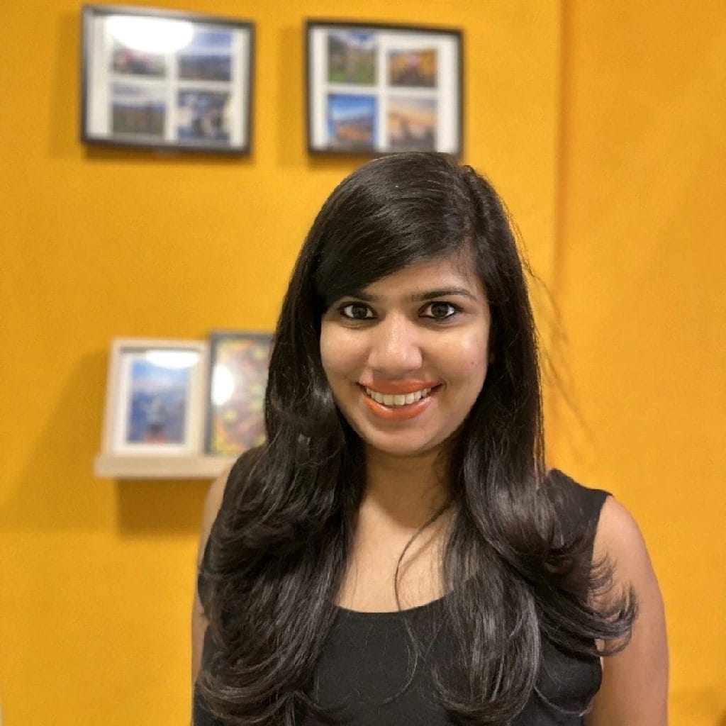 A woman, Prakriti Singh, smiling in front of a yellow wall with framed pictures.