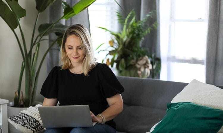 A woman sits on a gray sofa navigating her solo business landscape using a laptop in a cozy living room with a large plant and cushion accents.