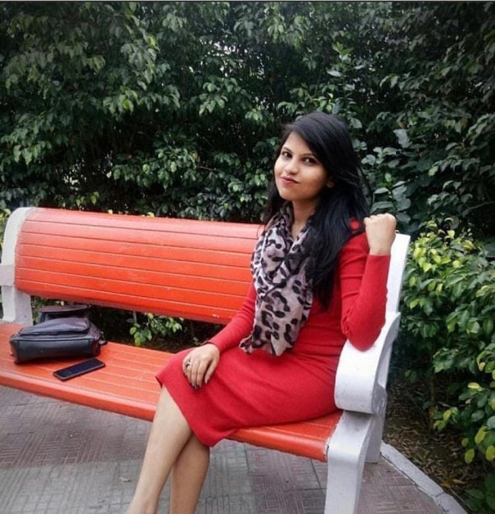 A young woman named Nisha Rawat in a red dress and leopard print scarf sits on an orange park bench, smiling at the camera.