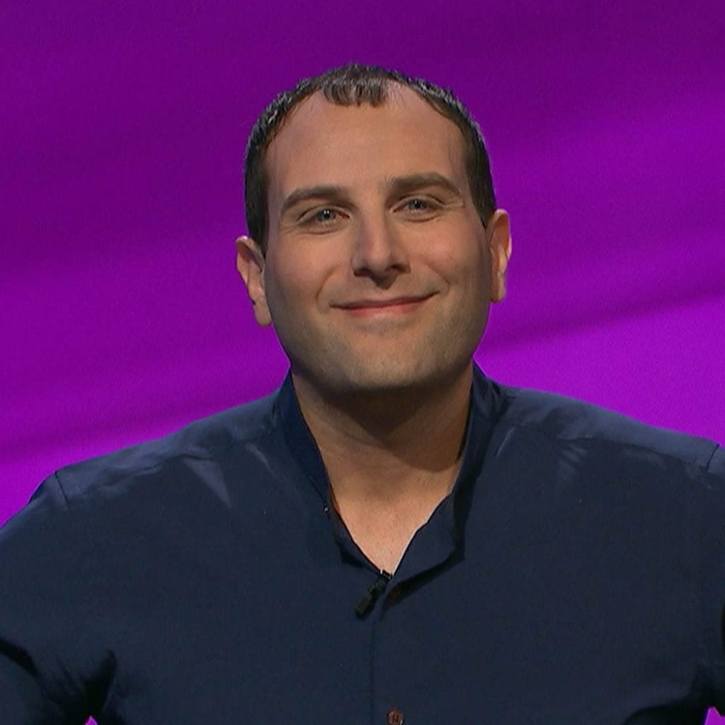 Shaun Gold, with a slight smile and dressed in a navy blue shirt, poses against a vivid purple background.