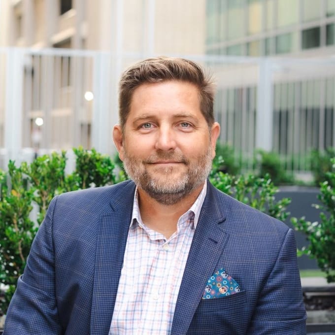 A man with a beard and short hair, resembling Jarie Bolander, is wearing a blue blazer and checkered shirt while sitting in an outdoor urban area with plants and buildings in the background.