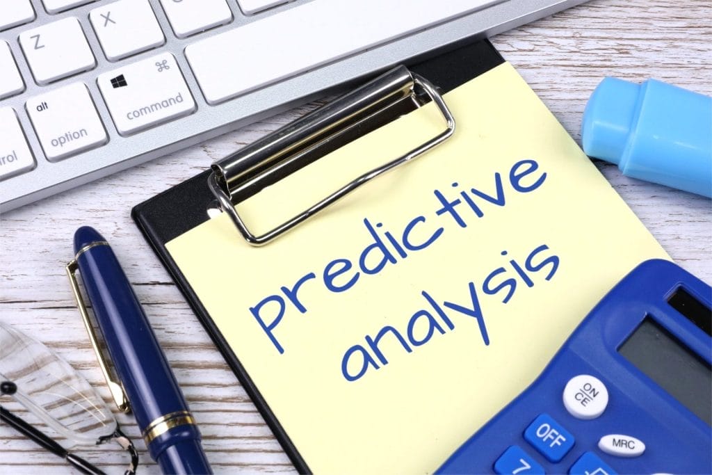 A clipboard with a yellow paper labeled "predictive analysis" next to a keyboard, pen, glasses, calculator, and blue marker on a desk&mdash;highlighting how AI is revolutionizing predictive analysis for small businesses.
