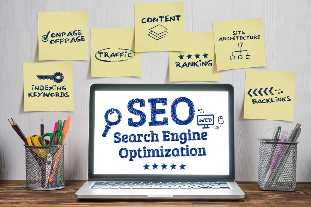 A laptop displaying "SEO Search Engine Optimization" sits on a desk, surrounded by sticky notes with SEO-related terms.